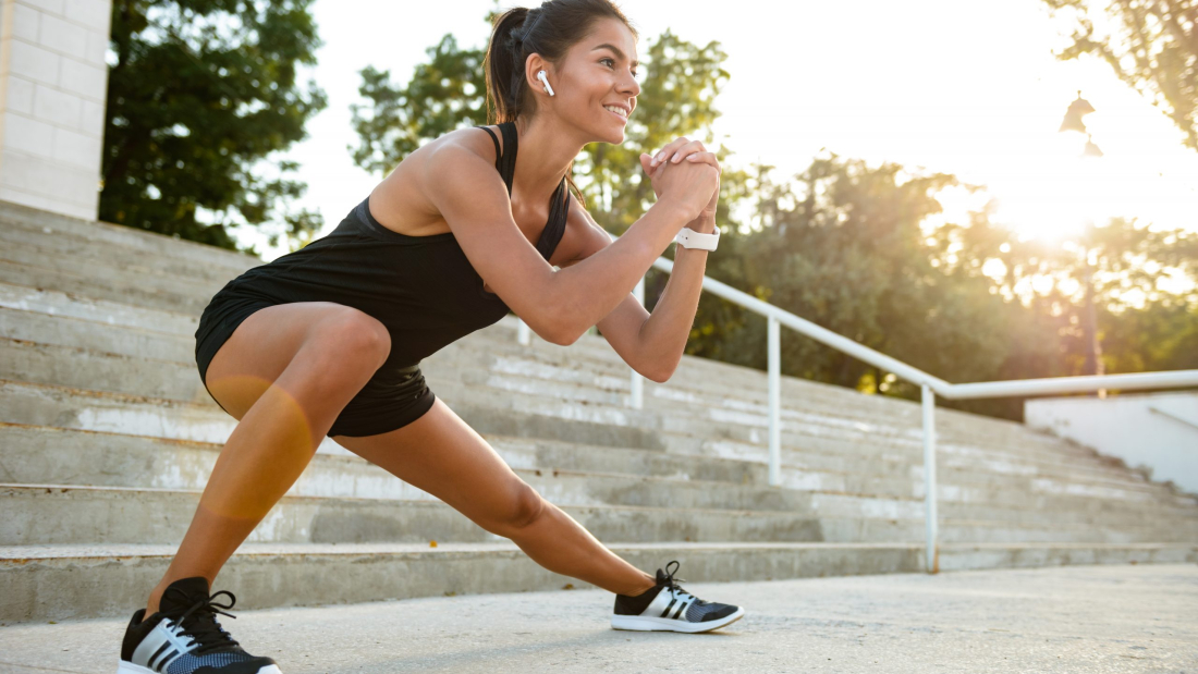 Portrait of a smiling fitness woman in earphones doing stretching exercises on stairs outdoors