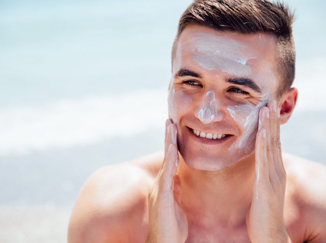 Cheerful man puts the lotion on face to protect a skin, on the beach