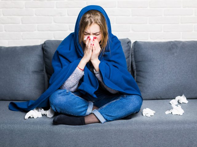 Sick woman with rheum and headache holding napkin, sitting on sofa with coveret and pills at home Sick woman with rheum and headache holding napkin, sitting on sofa with coveret and pills at home