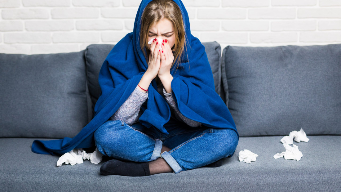 Mujer enferma con reuma y dolor de cabeza sosteniendo una servilleta, sentada en un sofá con cobertor y pastillas en su casa.