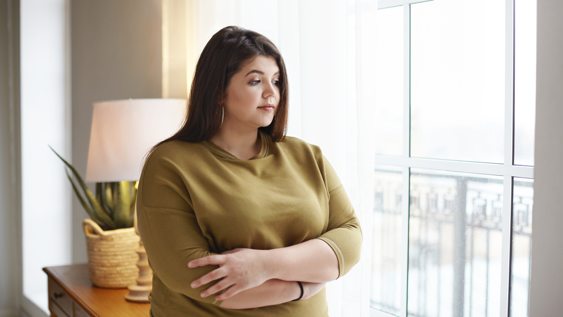 People, lifestyle and body positivity concept. Beautiful young brunette female with extra pounds standing by window at home, crossing arms on her chest, having thoughtful pensive look, pondering