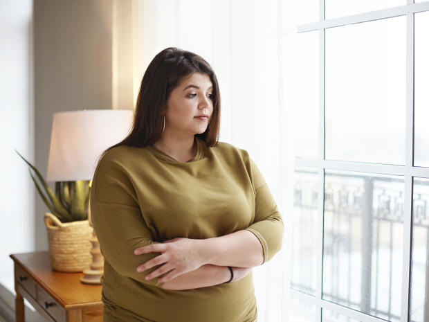 Concepto de personas, estilo de vida y positividad corporal. Hermosa joven morena con kilos de más de pie junto a la ventana en casa, cruzando los brazos sobre el pecho, teniendo mirada pensativa, reflexionando