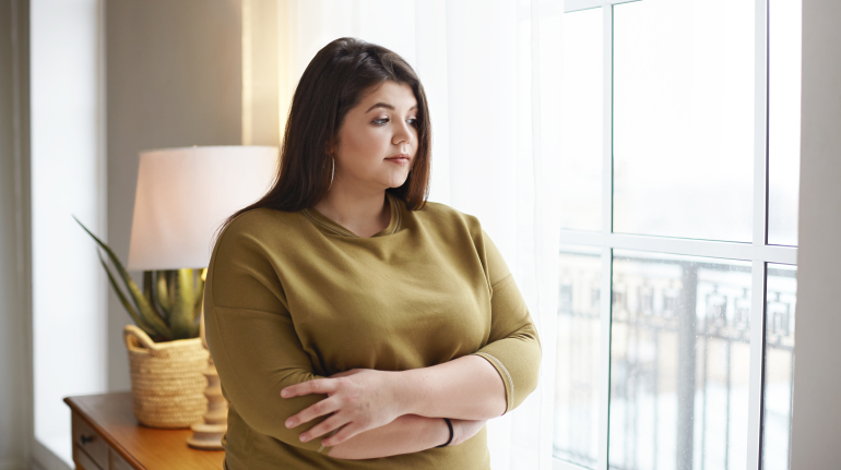 Concepto de personas, estilo de vida y positividad corporal. Hermosa joven morena con kilos de más de pie junto a la ventana en casa, cruzando los brazos sobre el pecho, teniendo mirada pensativa, reflexionando