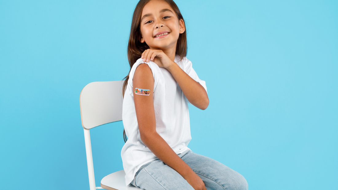 Vaccination Of Children. Happy Vaccinated Kid Girl Showing Arm With Adhesive Bandage After Vaccine Injection Sitting On Blue Studio Background. Kids And Covid-19 Prevention, Antiviral Immunization