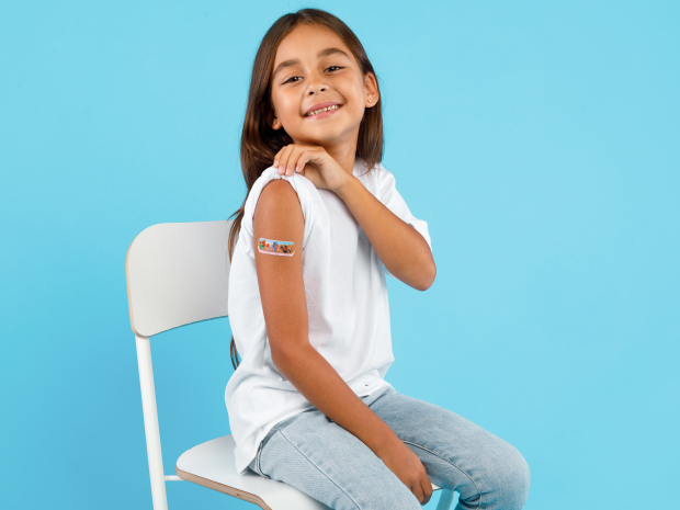 Vaccination Of Children. Happy Vaccinated Kid Girl Showing Arm With Adhesive Bandage After Vaccine Injection Sitting On Blue Studio Background. Kids And Covid-19 Prevention, Antiviral Immunization